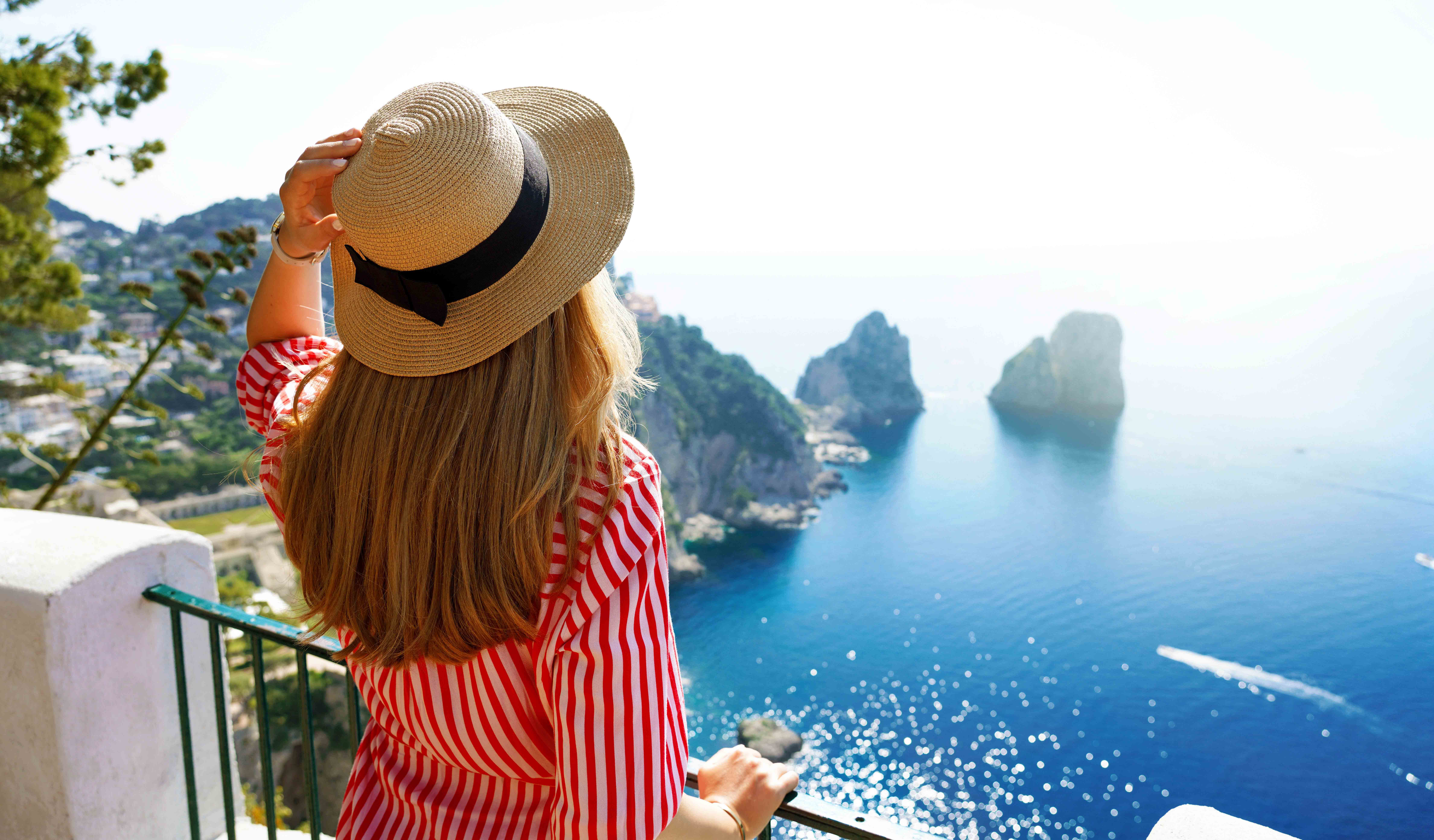 Beautiful young fashion woman with striped dress and hat in Capri Island with Faraglioni sea stack and blue crystalline water on the background, Capri, Italy